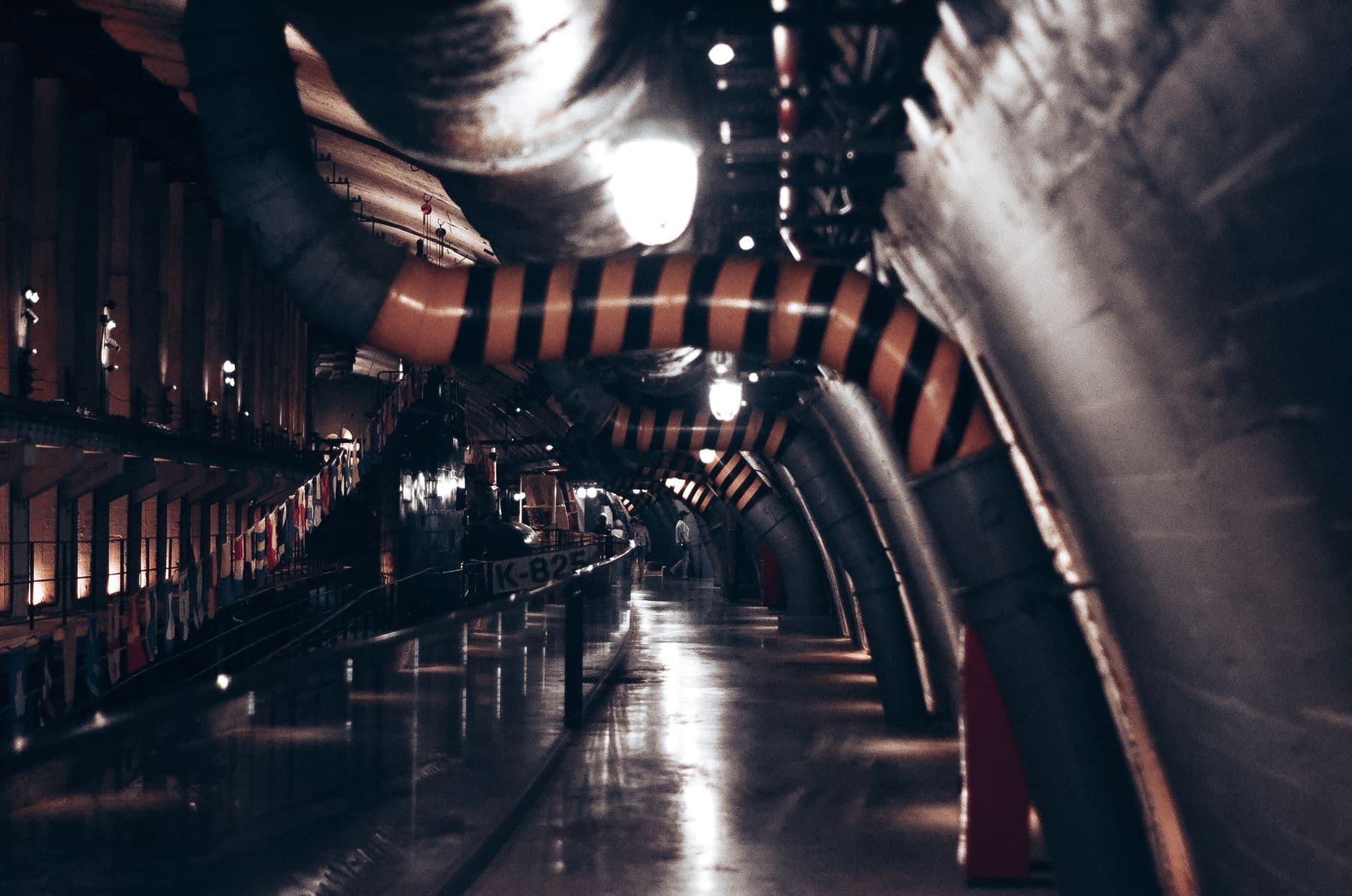 A dimly lit underground service tunnel lined with large pipes running into the distance